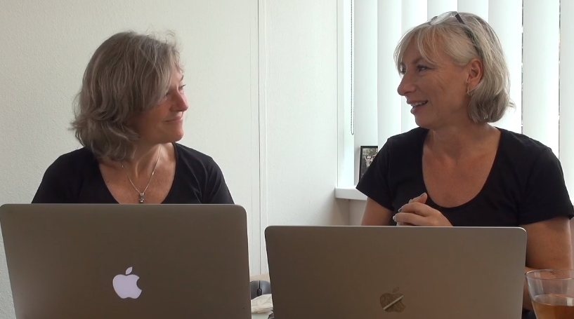 Foto van twee vrouwen met grijs haar die achter macbooks zitten en elkaar aankijken